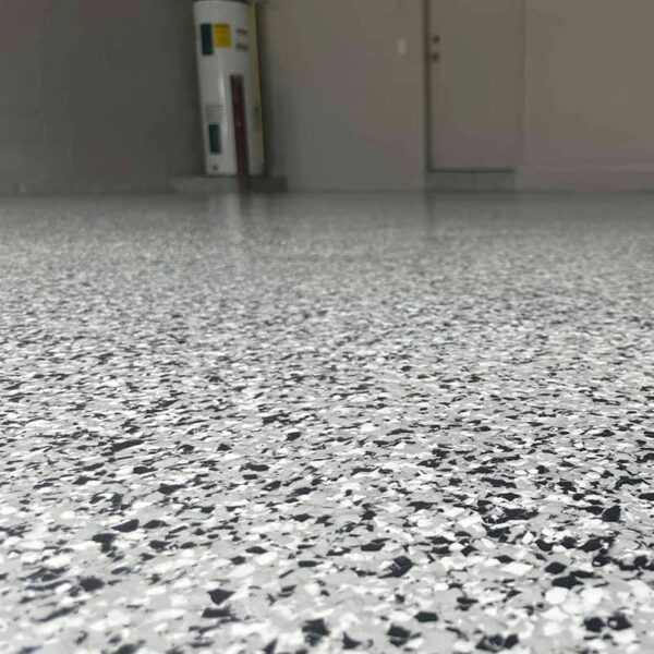 A close - up view of a garage floor exhibiting the application effect of polyurea coating, with a speckled pattern of white and black flakes, creating a durable and smooth surface.
