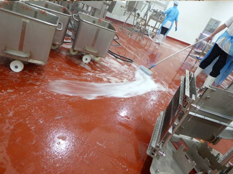 In a food processing workshop, a worker cleans the red chemical flame-retardant floor with a mop, creating white foam. Stainless steel material carts and processing equipment are visible, and another worker in protective clothing is in the background. The floor is flame-retardant, wear-resistant, corrosion-resistant, and easy to clean