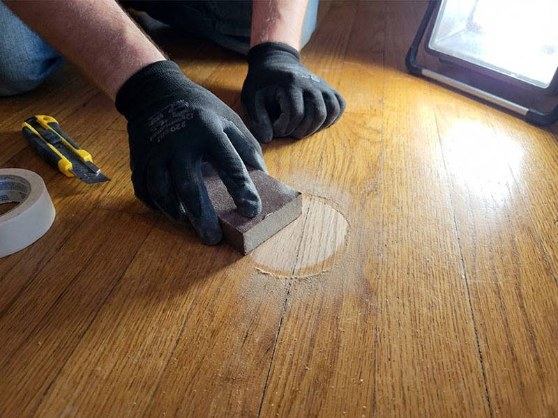 Hands using a small sanding block with fine-grit sandpaper to feather the edge of a small, peeling polyurethane patch on a wood floor.