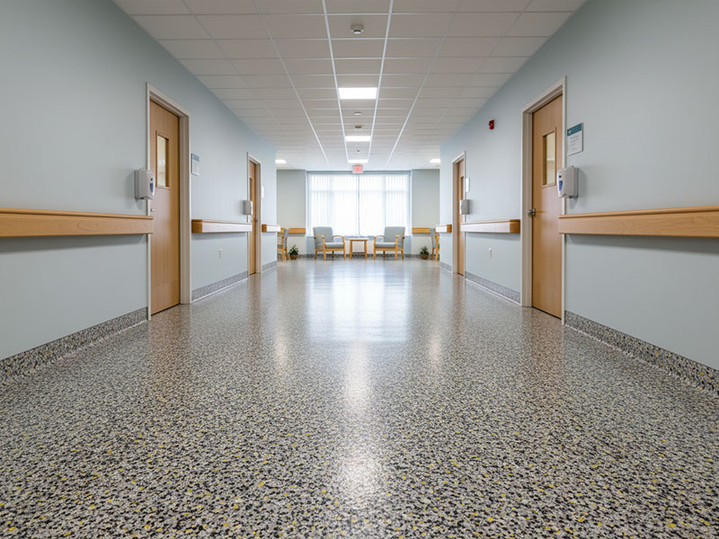 A clean, bright healthcare hallway featuring a durable four-layer flooring system with a decorative quartz aggregate and vinyl flake blend, showcasing a speckled grey and tan finish.