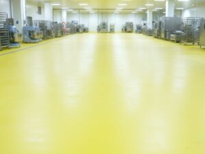 A wide-angle view of a clean industrial bakery facility featuring durable, bright yellow non-slip and food-safe resin flooring with commercial mixing equipment and racks lining the walls.