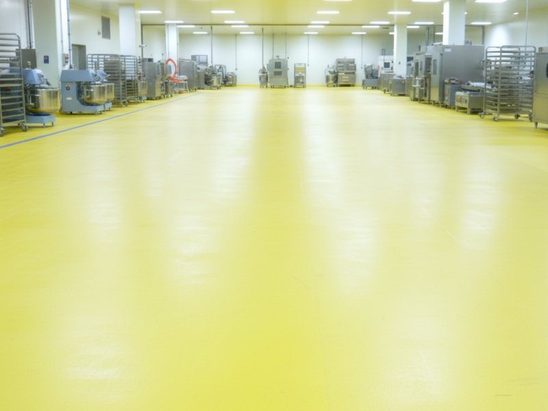 A wide-angle view of a clean industrial bakery facility featuring durable, bright yellow non-slip and food-safe resin flooring with commercial mixing equipment and racks lining the walls.