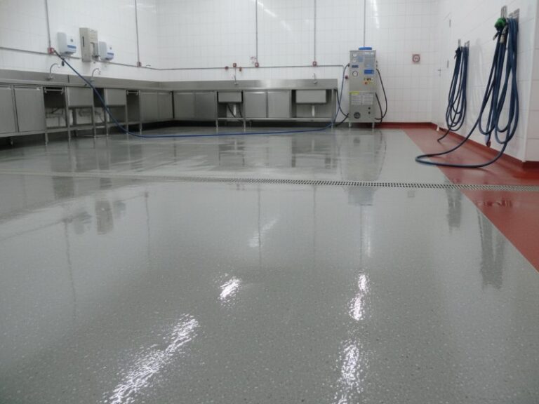A clean, seamless grey resin floor in a food processing facility washdown area with stainless steel sinks, white tile walls, and wall-mounted blue hoses.