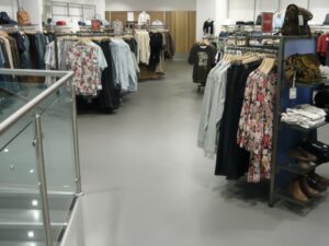 A wide view of a modern clothing retail store interior featuring racks of apparel, floral print shirts, and accessories like bags and shoes on display shelves.