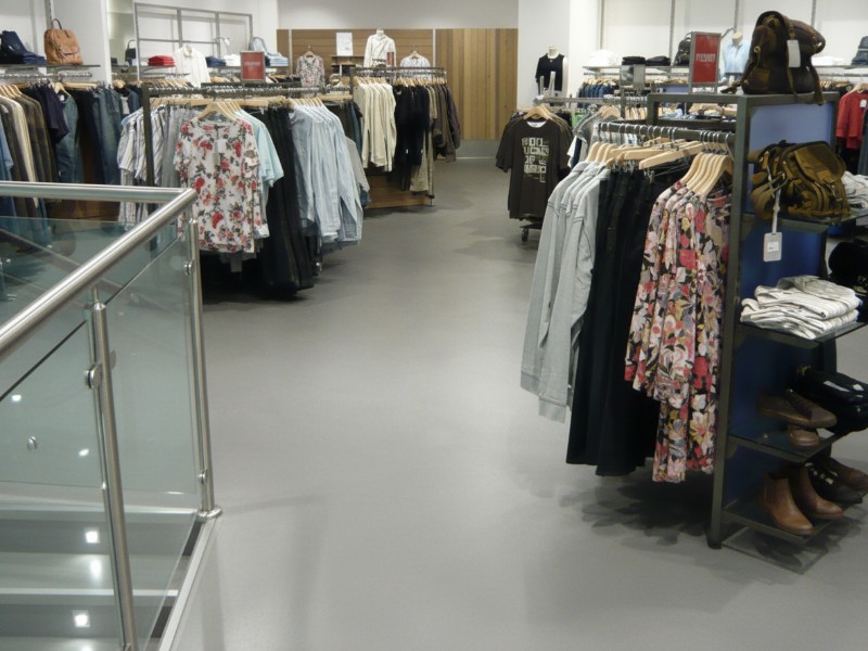 A wide view of a modern clothing retail store interior featuring racks of apparel, floral print shirts, and accessories like bags and shoes on display shelves.