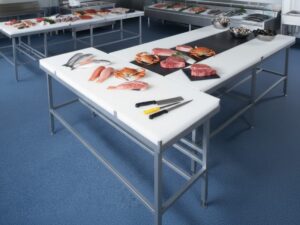 A professional commercial kitchen workspace featuring stainless steel tables with white polyethylene cutting board surfaces, displaying fresh seafood, raw steaks, and crabs for preparation.