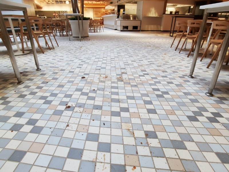 A wide-angle view of a checkered tile floor in a commercial food service area covered in visible food spills and stains, illustrating the maintenance challenges of industrial kitchen surfaces.