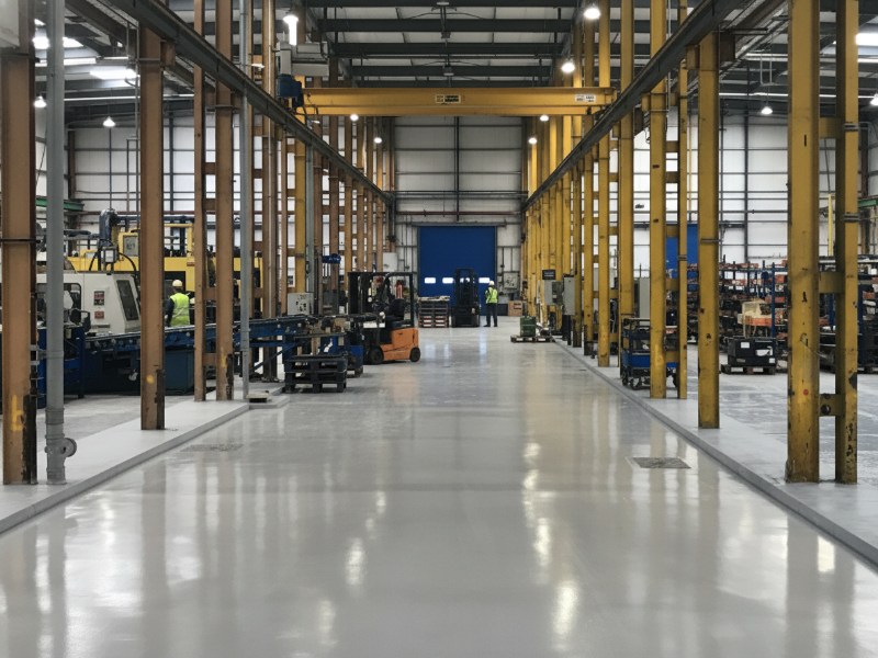 Light gray, highly reflective industrial floor spanning a vast manufacturing facility, flanked by yellow structural beams and machinery, with forklifts and workers present.