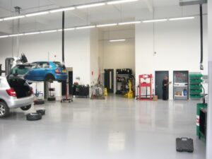 A clean, well-lit professional automobile service bay featuring a durable, light grey seamless garage floor coating with high light reflectivity, showing cars on lifts and a mechanic working in a repair shop environment.