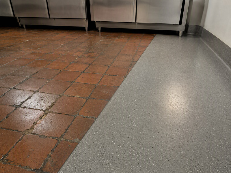 Split view of a commercial kitchen floor: worn clay tiles with dark eroded joints on one side, seamless resin with a coved edge on the other
