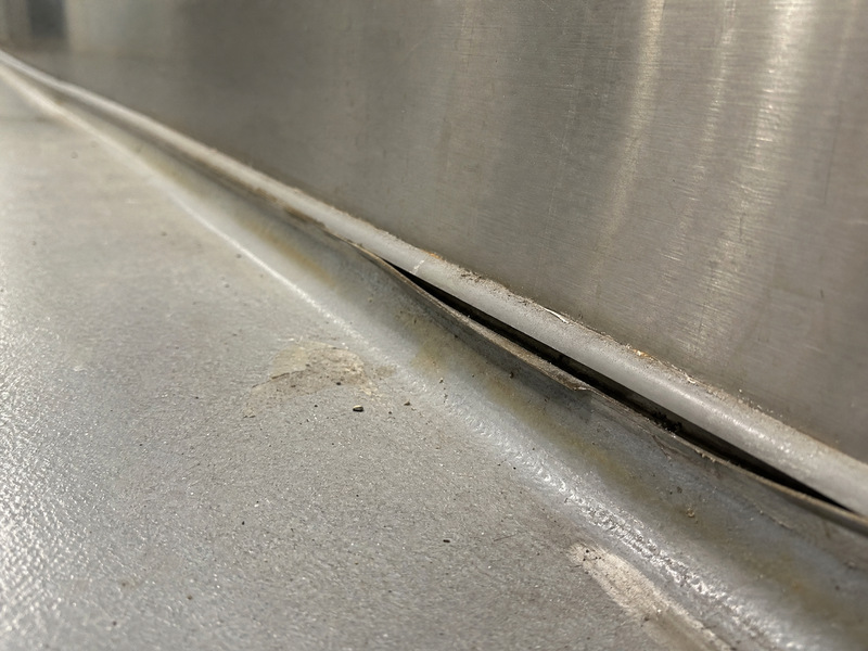 Failed sealant joint at a stainless steel plinth showing a gap at the cove head and soil trapped along the opened flank
